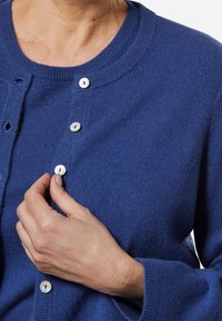 Blauer Wollcardigan mit rundem Ausschnitt, Knopfleiste mit kontrastierenden Knöpfen und gerippten Bündchen. Die Textur ist weich und leicht fusselig.