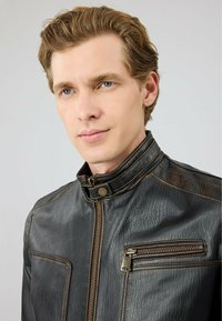 Zwarte leren jas met een hoge kraag, gestructureerde afwerking, twee ritszakken en bruine accenten langs de naden. Ritsluiting aan de voorkant.