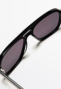 Schwarze rechteckige Sonnenbrille mit dunkel getönten Gläsern auf einer weißen Oberfläche, schräg gestellt, um den Rahmen und die Spiegelung der Gläser zu zeigen.