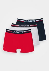 Drei Paar Herrenboxershorts in Rot, Grau und Marineblau. Elastische Bündchen mit Lacoste-Logo und farbigen Streifen. Glatte Stofftextur.