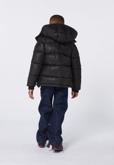 Giacca a vento nera con cappuccio, design trapuntato, texture lucida e un motivo sottile. Indossata con jeans in denim blu e scarpe colorate.