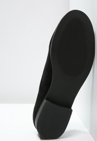 Chaussure en suede noir avec un bout rond et une semelle en caoutchouc texturée basse, présentant des lignes discrètes et une finition mate.