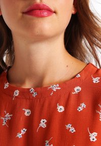 Roestkleurige blouse met een ronde halslijn, voorzien van een bloemmotief in wit en lichtblauw. Zachte, gestructureerde stof.