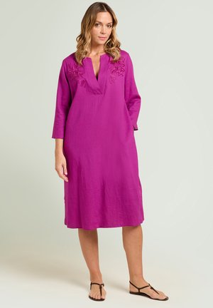 Femme portant une robe magenta longueur genou avec manches longues, broderie florale sur les épaules et sandales plates noires, debout sur un fond neutre.