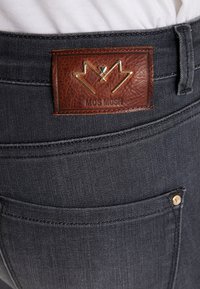 Närbild av mörkgrå denimjeansficka med brun läderetikett från Mos Mosh och mässingsnipplar i fickans hörn.