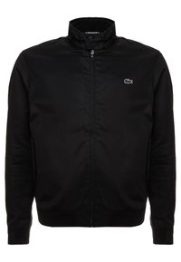 Svart zip-up jacka med ståkrage, knappstängning vid halsen, sidofickor och liten vit krokodillogo på vänster bröst.