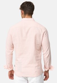 Hellrosa Langarmhemd mit weißen vertikalen Streifen, einer Rückenpasse und knöpfbaren Manschetten. Hergestellt aus einem weichen, strukturierten Stoff.