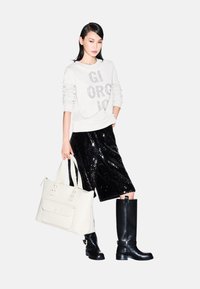 Sudadera blanca con "GI ORG IO" en purpurina, falda midi negra con lentejuelas, botas altas negras y una bolsa tote blanca con asa y bolsillo frontal.