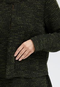 Czarna i zielona kurtka o fakturze, luźny krój, prosty dekolt, długie rękawy oraz widoczne detale kieszeni.