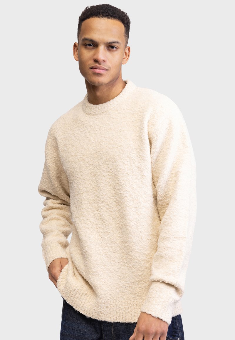 Beige, textured sweater with a round neckline and ribbed cuffs. Relaxed fit, featuring a soft, fuzzy material, paired with dark denim jeans.