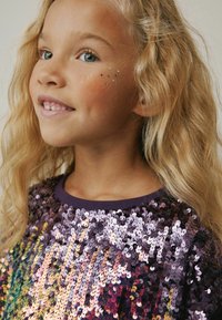 Lilla paillet-top med et farverigt gradientdesign. Lys bølget hår, blå øjne og stjerneformede glimmer accenter på kinderne.