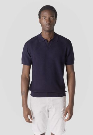 Joven de pie, vestido con una camiseta polo de manga corta azul marino con textura y pantalones cortos blancos desgastados, sobre un fondo liso y claro.