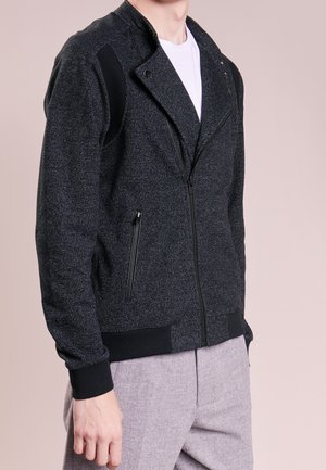 Zwarte zip-up jas van textuurstof, met een biker-stijl kraag, zijzakken en zwarte accenten op de schouders en manchetten.