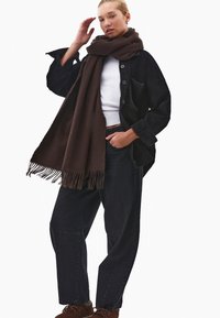 Przesadzona czarna kurtka koszulowa z kordurowej tkaniny, biała koszulka, ciemne dżinsy, owinięta ciemno-brązowym wełnianym szalem z frędzlami. Brązowe buty typu lace-up.