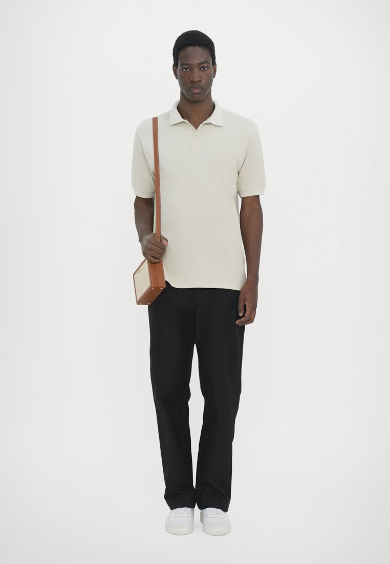 Polo beige, pantalons noirs, baskets blanches et un sac en bandoulière marron. Le modèle se tient debout contre un fond blanc.