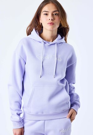 Giovane donna con una felpa con cappuccio viola chiaro e pantaloni della tuta abbinati con il logo "Project X Paris", in piedi contro uno sfondo bianco uniforme.