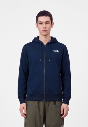 Joven con sudadera con capucha azul marino con cremallera y logo de The North Face, y pantalones verde oliva, de pie frente a un fondo blanco liso.