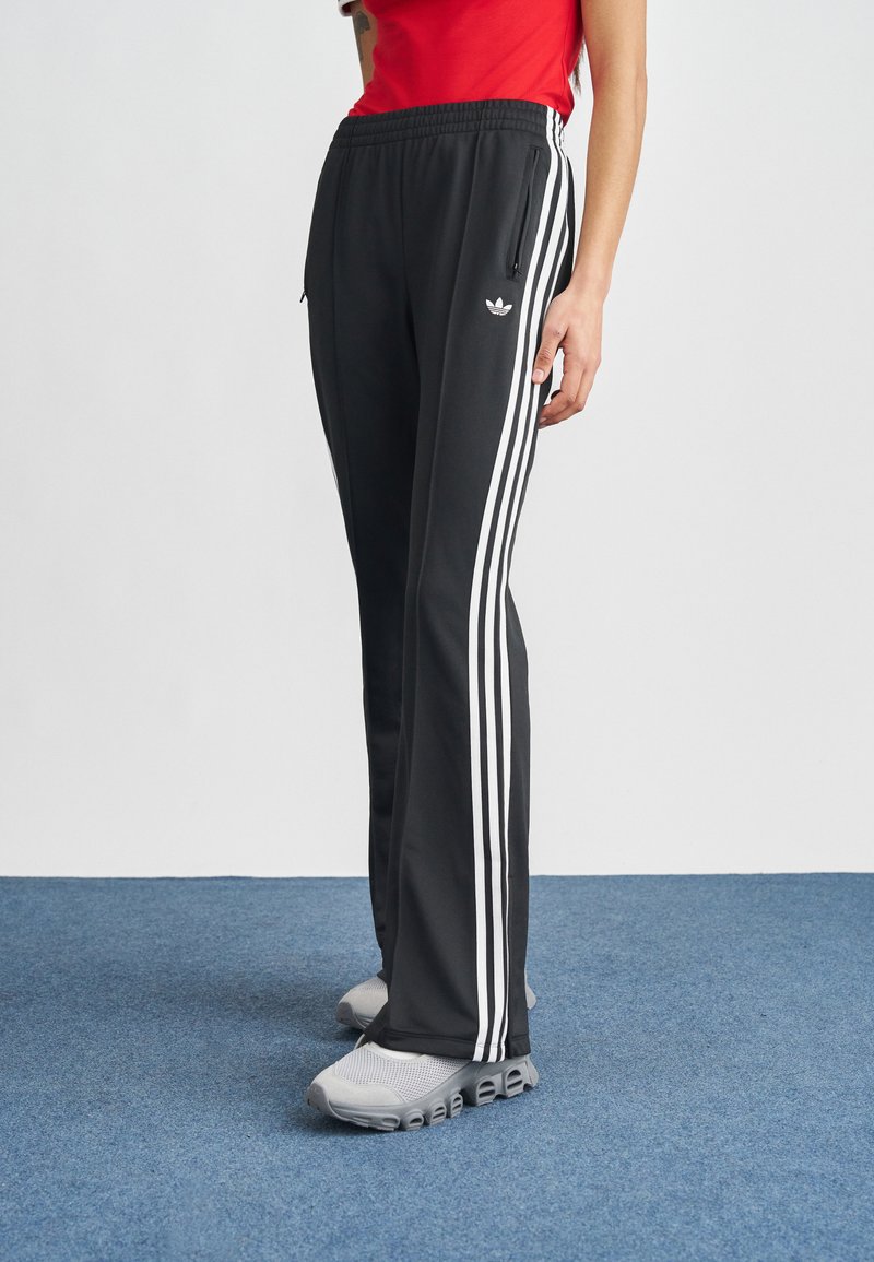 Pantalones deportivos negros con franjas blancas a los lados, con cintura ajustable con cordón y bolsillos con cremallera, combinados con zapatillas grises.