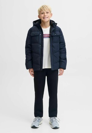 Marinblå puffjacka med huva, med en kviltad design och fyra framfickor, bärs över en vit t-shirt och svarta byxor, tillsammans med sneakers.