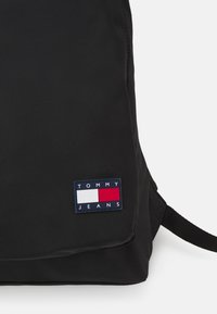 Svart ryggsäck med en rektangulär Tommy Jeans-logotyp i röd, vit och marinblå. Slät tyg med justerbara axelremmar.