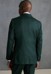 Chaqueta de traje ajustada de color verde oscuro con una textura suave, abertura sencilla en la parte posterior y un brillo sutil. Presenta botones negros y una camiseta blanca por debajo.