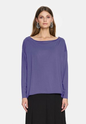 Kobieta w długim fioletowym topie z szerokim dekoltem i czarnej spódnicy, ozdobiona dużymi srebrnymi kolczykami w kształcie kwiatów, stojąca na tle białym.