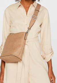 Tan crossbody-taske med en struktureret form, glat kunstlædermateriale og en kontrasterende sort kant. Justerbar rem med metalbeslag.