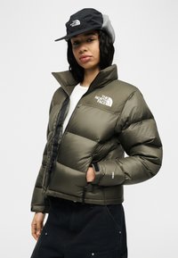 Άτομο που φοράει μπουφάν North Face σε ελιά πράσινο χρώμα με επένδυση και μαύρο καπέλο North Face με καλύμματα αυτιών, στέκεται με το ένα χέρι στην τσέπη του μπουφάν.