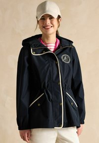 Jūras spēku ūdensnecaurlaidīga jaka ar kapuci, regulējamu auklu jostas vietā, baltām maliņām un zelta pogām. Zem tās redzama balta svītrainā krekla.