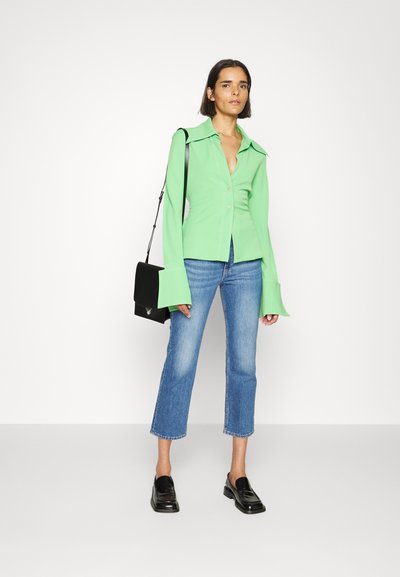 Grünes Hemd mit breiten Manschetten, kombiniert mit hellblauen, knöchellangen Jeans und schwarzen Loafers. Minimalistische schwarze Tasche mit Klappe.