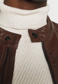 Hnědá kožená bunda s límcem na cvočky, vybavená černým zipem a knoflíky. Pod ní je vidět krémový žebrovaný rolák.