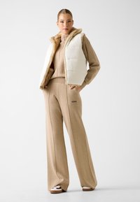 Gewatteerd vest in het wit met een beige nepbontvoering, gedragen over een bijpassende beige trui en wijde broek met het "GUESS"-logo.