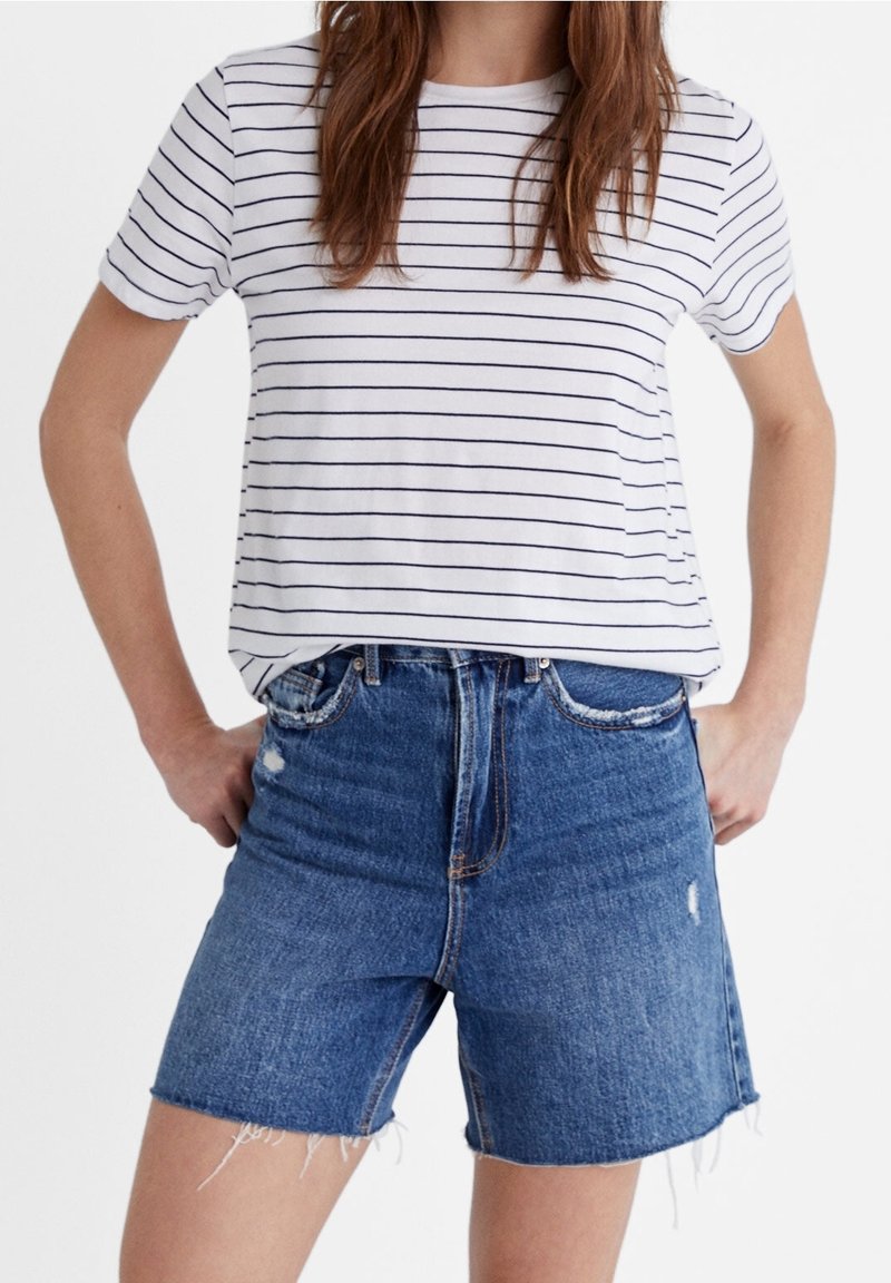 Femme portant un haut blanc à manches courtes avec de fines rayures noires horizontales et un short en denim bleu taille haute avec des ourlets effilochés.