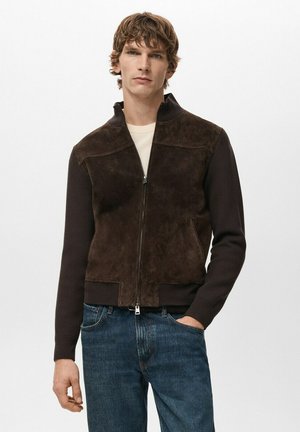 Joven con una chaqueta de ante marrón oscuro con cremallera y vaqueros azules, de pie con una mano en el bolsillo frente a un fondo liso.