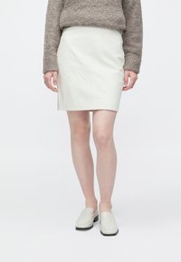 Bela usnjena mini krilo z gladko teksturo. Združeno z mehkim bež puloverjem in svetlimi natikači, ki predstavljajo monokromatsko paleto.