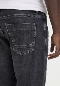 Jeans de denim negro con un acabado texturizado, bolsillos traseros estándar con costuras visibles y una pequeña etiqueta de logo cerca de la cinturilla.