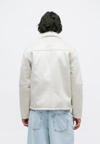Giacca in suede beige chiaro con colletto in pelliccia, spalle strutturate e cuciture a contrasto, indossata sopra jeans di denim azzurro chiaro.