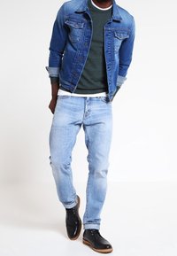 Mężczyzna w niebieskiej dżinsowej kurtce na ciemnozielonym swetrze, w jasnoniebieskich dżinsach i czarnych skórzanych butach, idzie na tle białym.