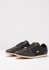 Svarta träningsskor med en blandning av texturerade material, guldöglor och en vit gummisula. Inkluderar en subtil logotyp på sidan.