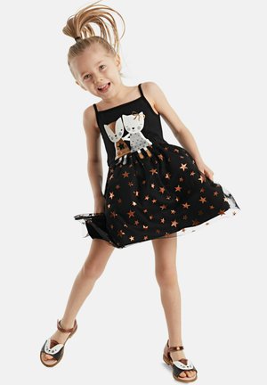 Schwarzes Kleid mit einem taillierten Oberteil, das mit Tiermotiven verziert ist; der Rock ist mit metallischen Sternen geschmückt. Kombiniert mit braunen und schwarzen Sandalen.
