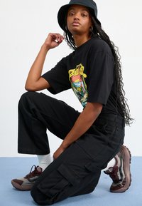T-shirt grafica nera con design colorato, pantaloni cargo neri e sneakers grigio-marroni con dettagli rossi. Il modello è in posa su un ginocchio.