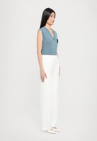 Haut en tricot sans manches de couleur bleu clair avec un col en V et des accents noirs, associé à un pantalon large plissé blanc, complété par des chaussures à enfiler crème.