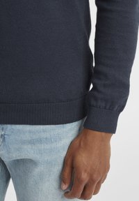Maglione a maglia blu navy con orlo e polsini a costine, indossato su jeans in denim azzurro, che mettono in mostra una texture del tessuto liscia.