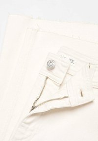 Jeans en denim beige avec fermeture éclair argentée et détail de bouton, dotés d'une étiquette avec des informations sur la marque fixée à la taille.