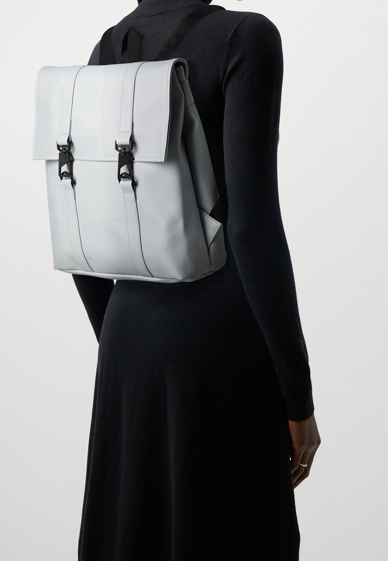 Personne portant un sac à dos rectangulaire gris clair avec des sangles à boucle noire sur une robe noire à manches longues sur un fond uni.