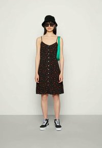 Vestido preto com pequeno padrão floral vermelho, alças finas, fecho de botões à frente, barra larga, combinado com tênis pretos e uma bolsa verde.