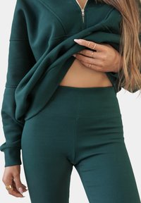 Donkergroene ritstrui met ribbelstructuur, gecombineerd met bijpassende ribbel hoge taille leggings. Zichtbare buik met handen die de stof vasthouden.