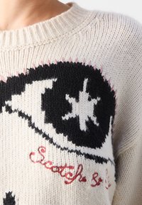 Pull en tricot crème avec un motif graphique noir, présentant un motif d'étoile et des accents roses. Texte « Scotch & » en détail de broderie rouge.