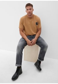 Lichtbruine katoenen t-shirt met een zwart grafisch ontwerp op de borst, gecombineerd met vervaagde grijze spijkerbroek en zwarte enkellaarsjes met rode accenten.