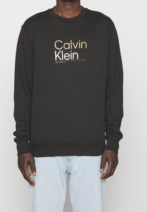 Osoba ubrana w czarną bluzę Calvin Klein z logo w złotym i białym kolorze, w połączeniu z jasnoniebieskimi dżinsami, pokazana od szyi do bioder.